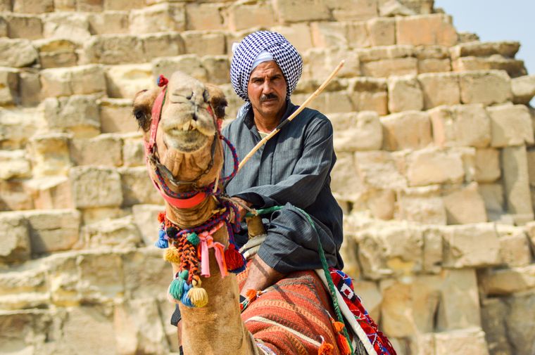 A camel handler in a checked headscarf on a decorated camel in front of ancient stone blocks