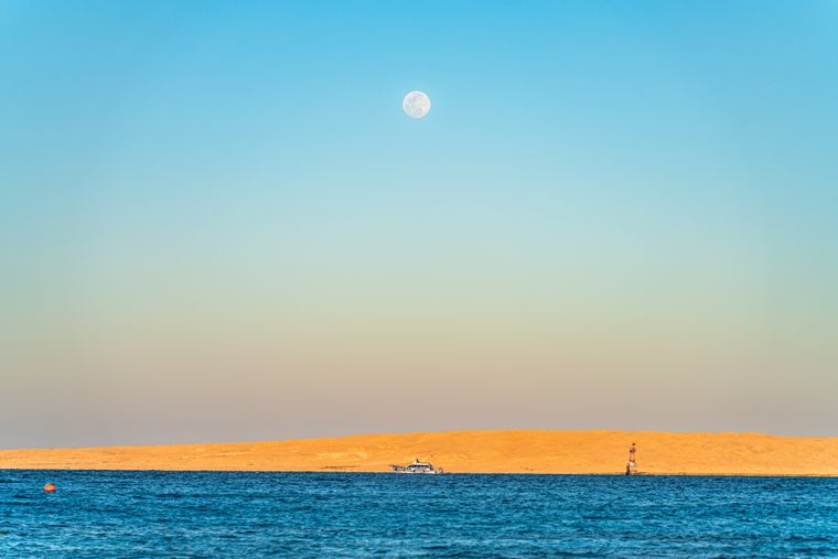 A full moon above a sandy island and a small boat on the deep blue Red Sea